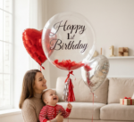 Прозрачный шар Bubble с черной надписью Happy 1st Birthday, красными перьями внутри и фольгированными сердцами в интерьере.