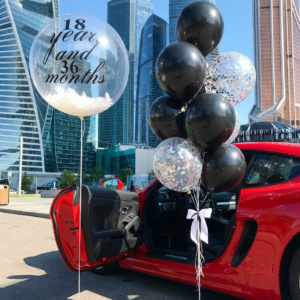 Прозрачный шар Bubble с перьями и надписью 18 years and 36 months рядом с связкой черных шаров с конфетти на фоне небоскребов.