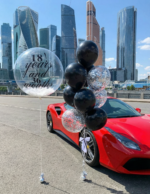 Прозрачный шар Bubble с перьями и надписью 18 years and 36 months рядом с связкой черных шаров с конфетти на фоне небоскребов.