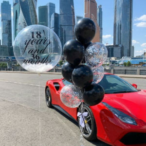 Прозрачный шар Bubble с перьями и надписью 18 years and 36 months рядом с связкой черных шаров с конфетти на фоне небоскребов.